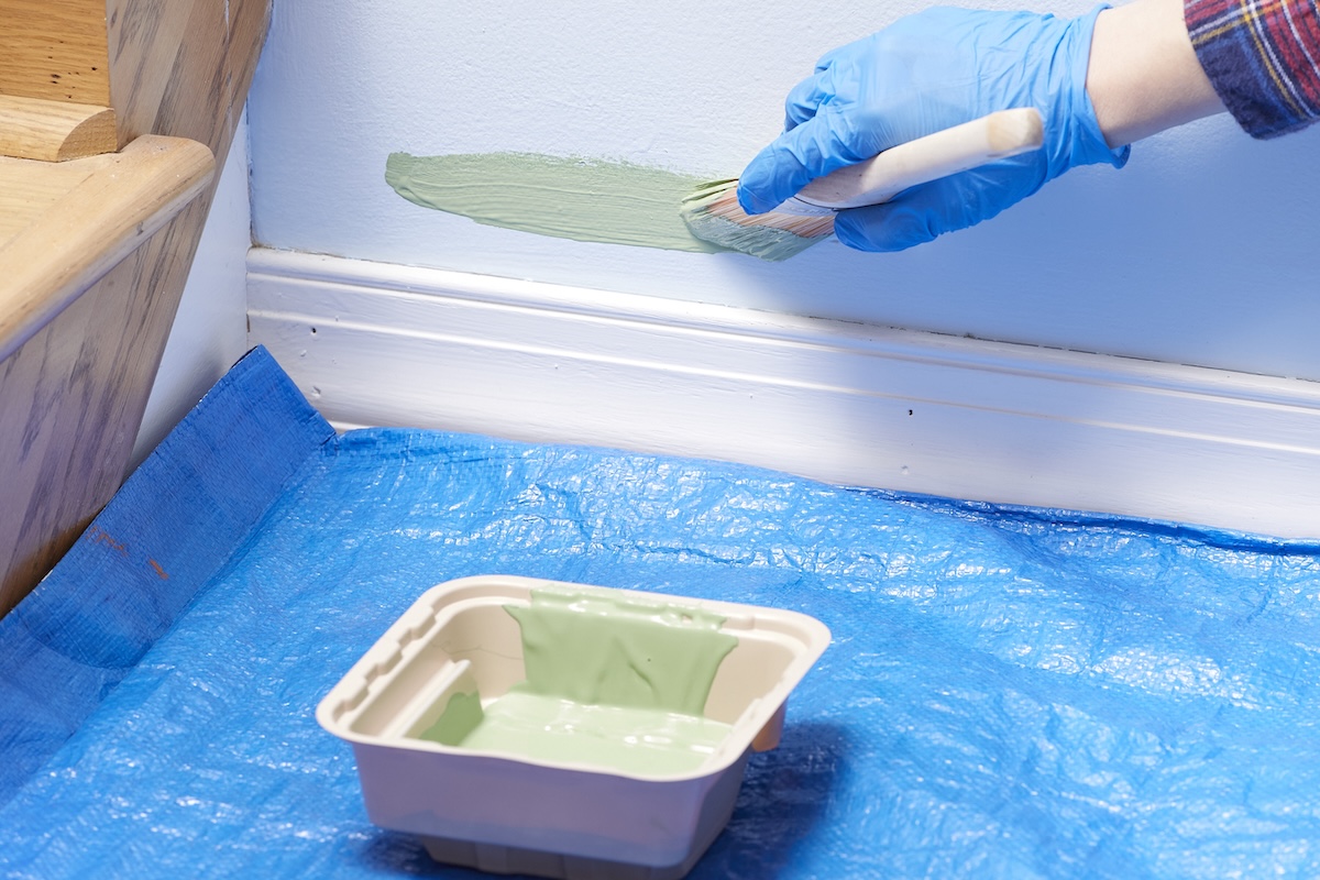 Person wearing a blue glove paints green paint on a wall about an inch above the baseboard.