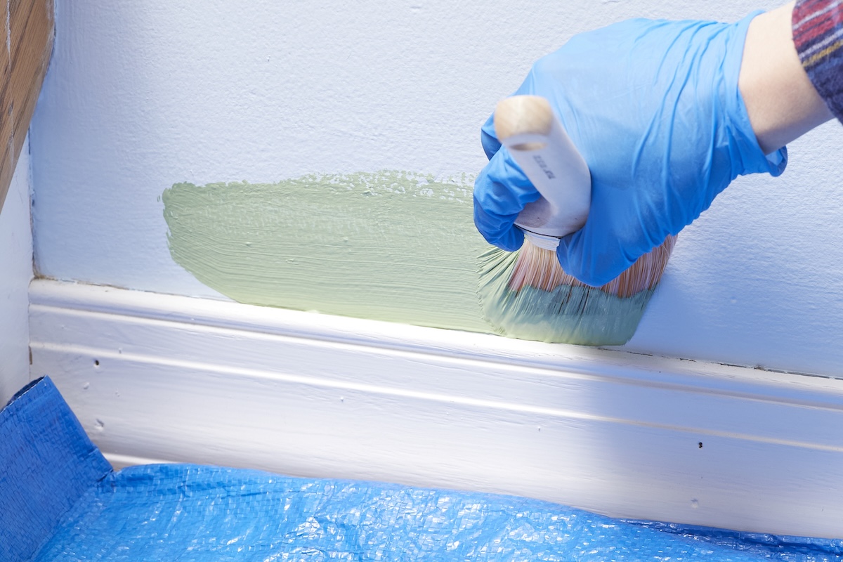 Person wearing a blue nitrile glove paints light green paint on a wall very close to a baseboard.