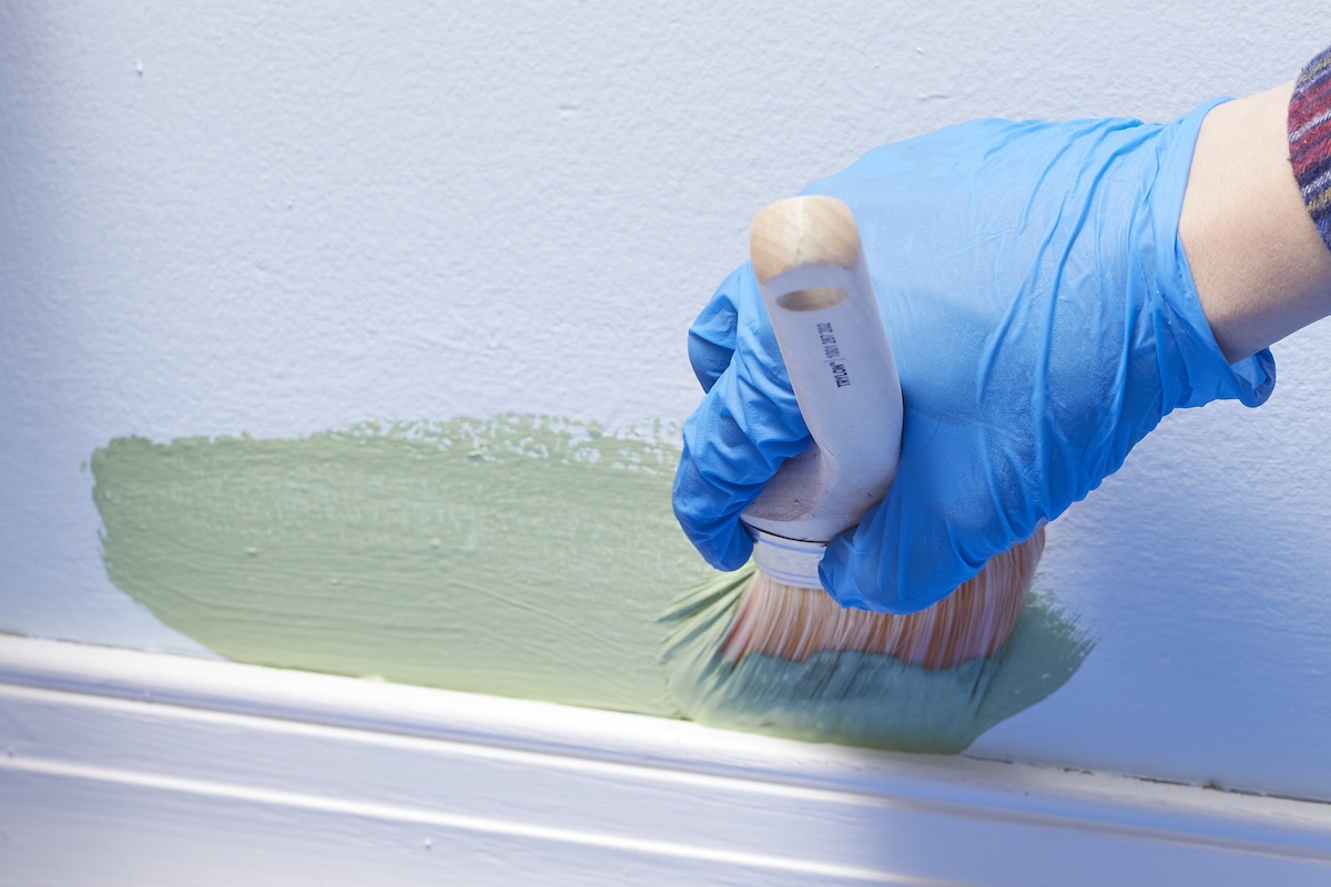 Person wearing blue nitrile glove uses paint brush to cut in light green paint near a baseboard.