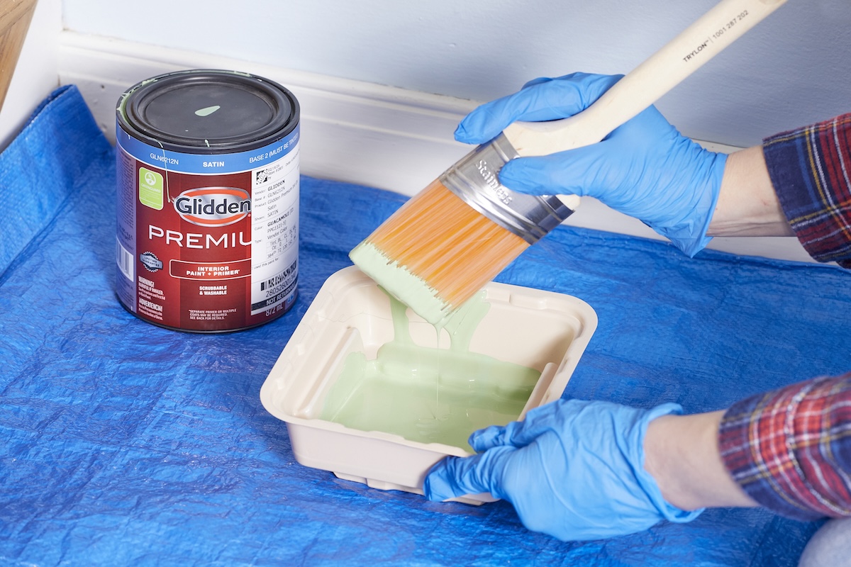 Person dips paintbrush into a paint trough filled with light green paint.