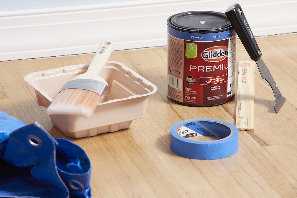 Can of paint, paintbrush, tarp, and other painting materials on a wood floor.