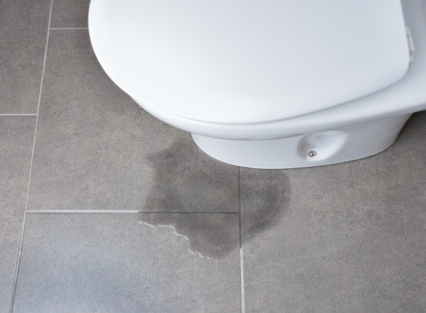 A puddle of water is leaking from the bottom of a toilet onto the grey tiled floor.