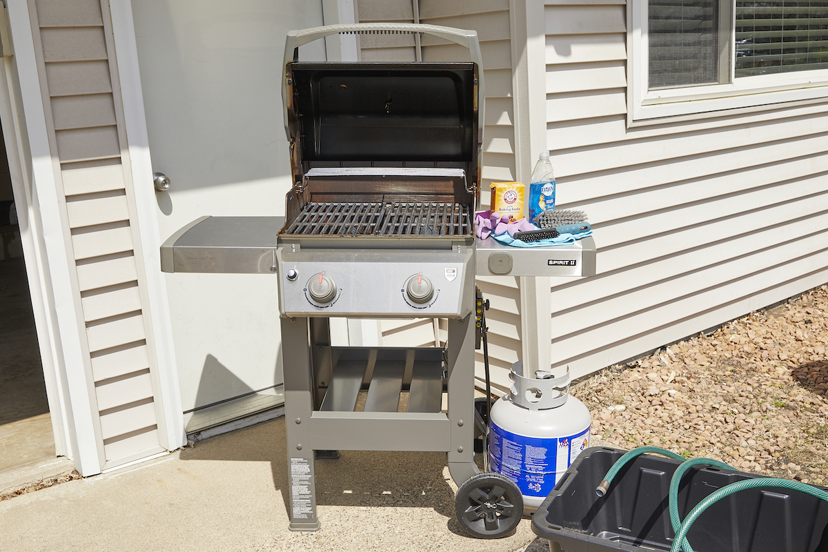 A gas grill with the top open, surrounded by materials and tools for cleaning it (including a plastic bin, garden hose, dish soap and more).