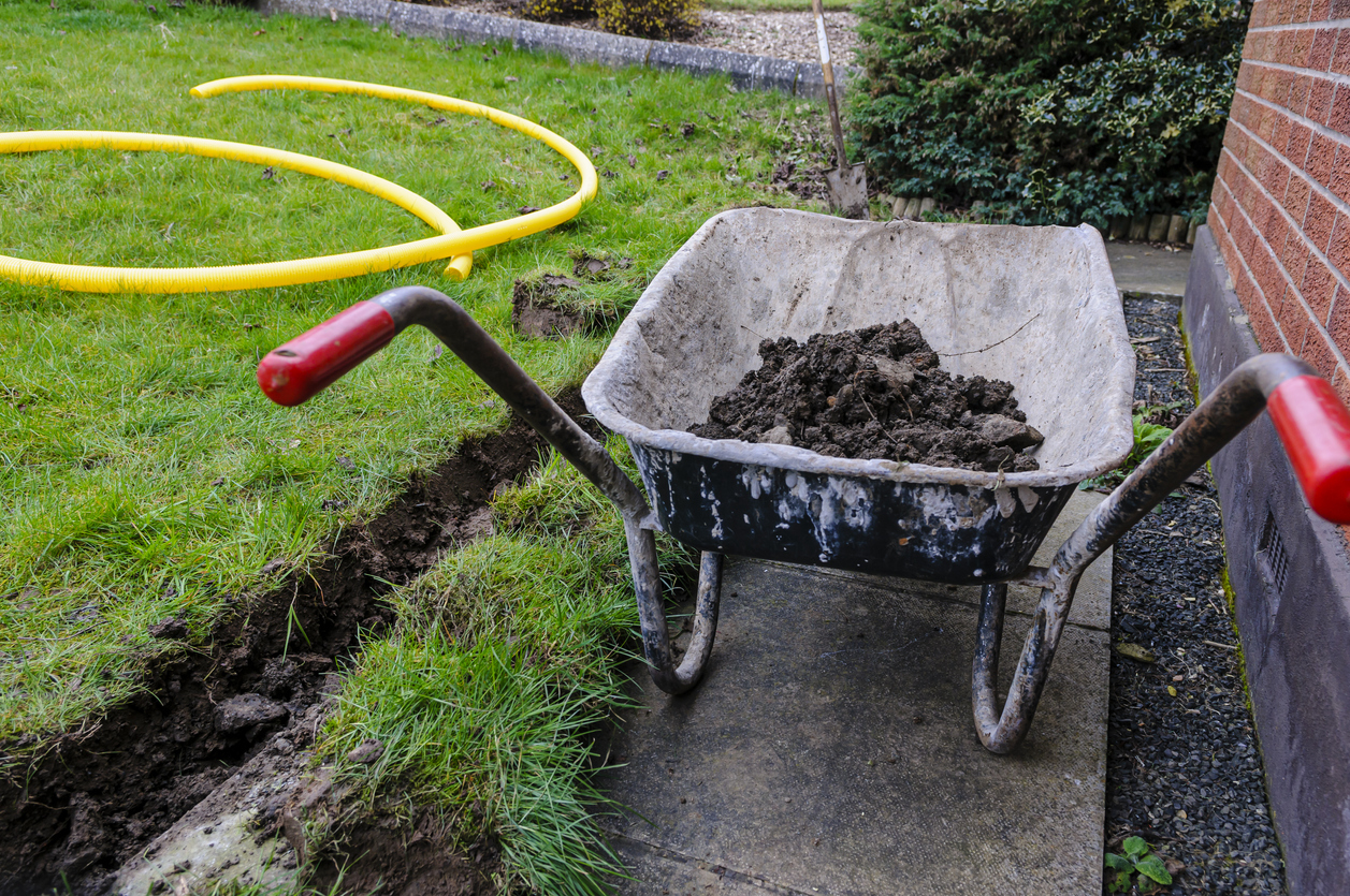 Wheelbarrow full of soil after a trench has been dug at the bottom of a lawn to install a drainage channel.