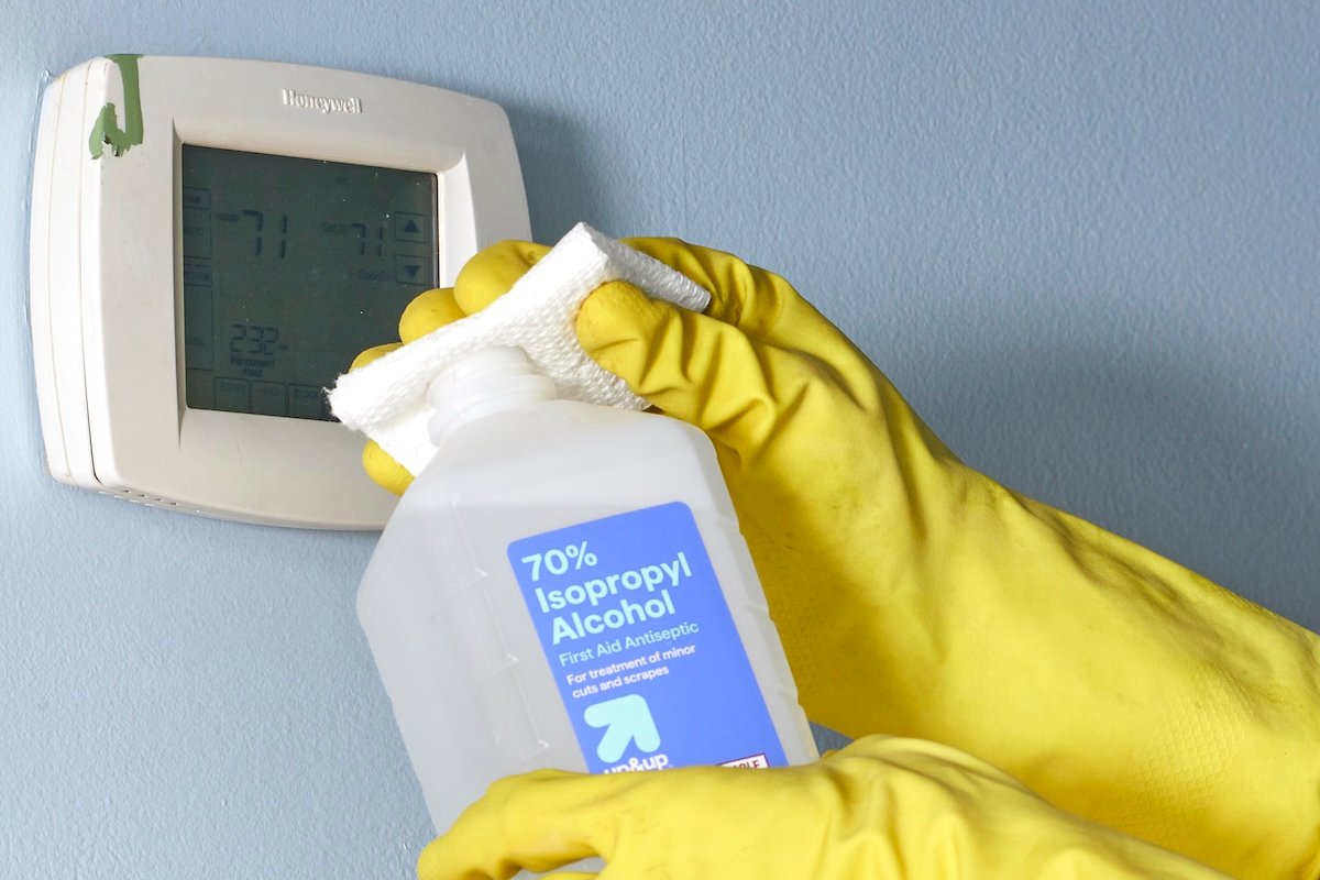 Woman wearing yellow rubber gloves dabs rubbing alcohol on a paper towel, ready to remove green paint drips on a wall thermostat.
