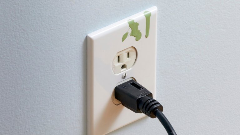 Splatter of green paint on a white wall electrical outlet cover; a black cord is plugged into the outlet.