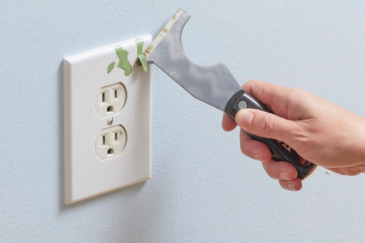 Woman uses a paint scraper to lift green paint splatters on a white electrical outlet cover.