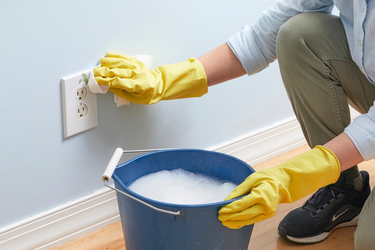Woman wearing yellow rubber gloves washes an electrical outlet, a bucket of soapy water near her other hand.