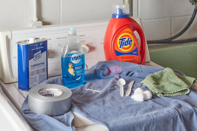 On top of a top-load washing machine, a paint-spattered shirt and the materials needed to remove paint from the shirt.