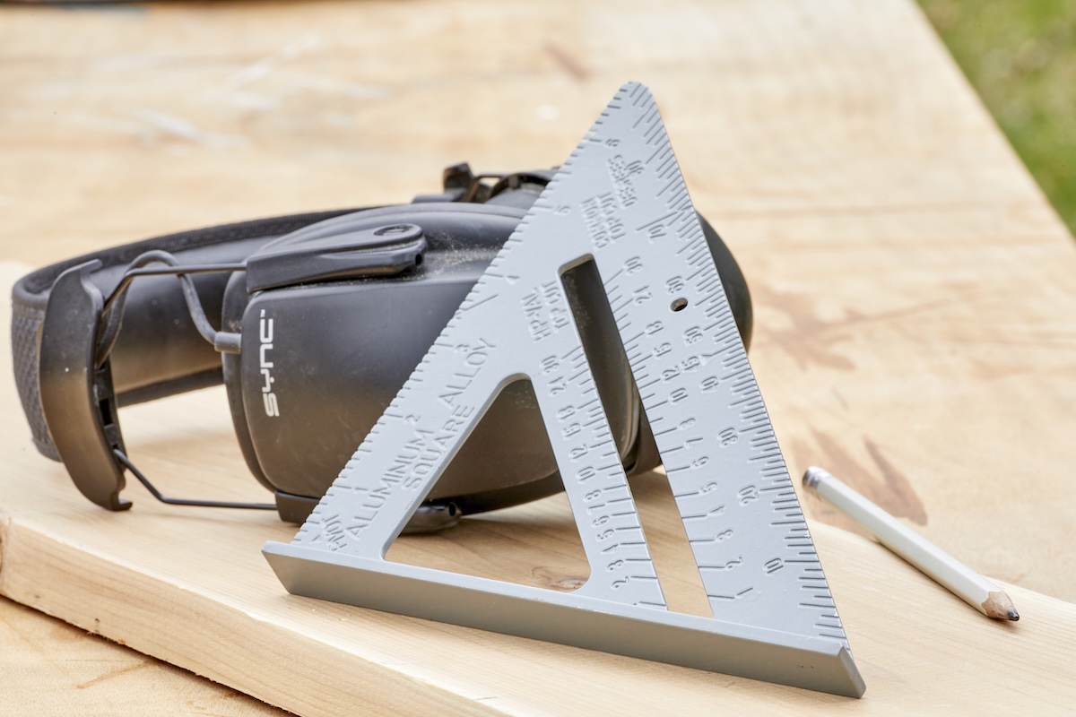 A speed square is propped up against ear protecting headphones, a pencil nearby.