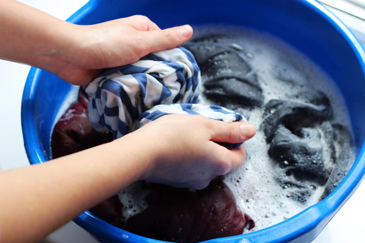 Person hand-washing soapy clothes in a blue bucket.