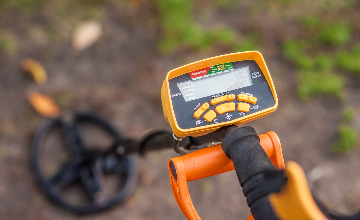 A metal detector in yellow and orange housing pointed toward the soil.