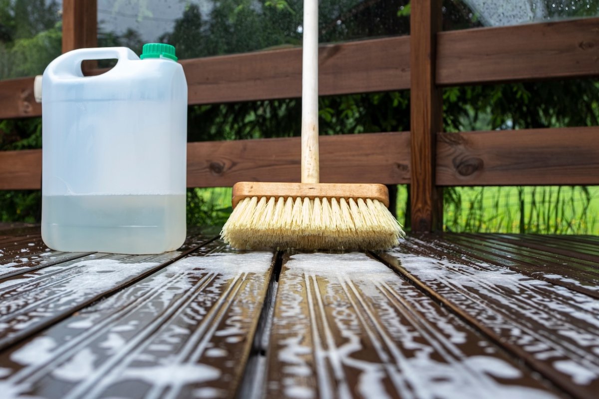 A deck scrub brush and a plastic container of cleaning solution sit on a soapy wood deck.