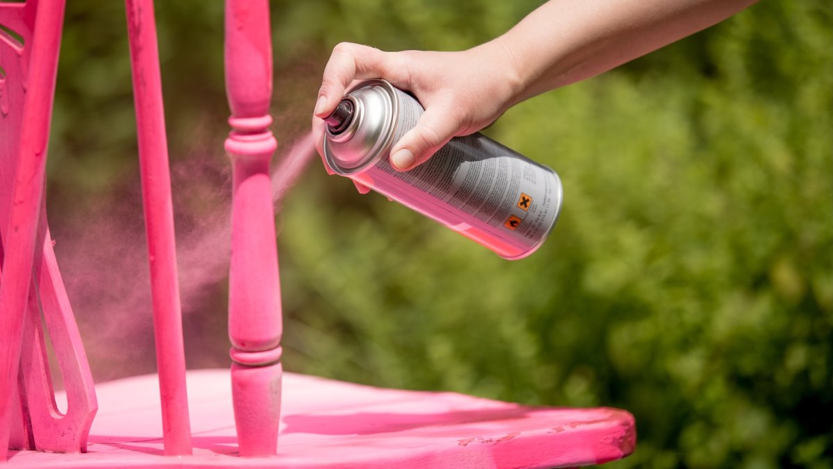 Person spray paints a spindle chair hot pink, outdoor greenery in the background.