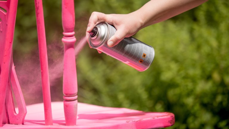 Person spray paints a spindle chair hot pink, outdoor greenery in the background.