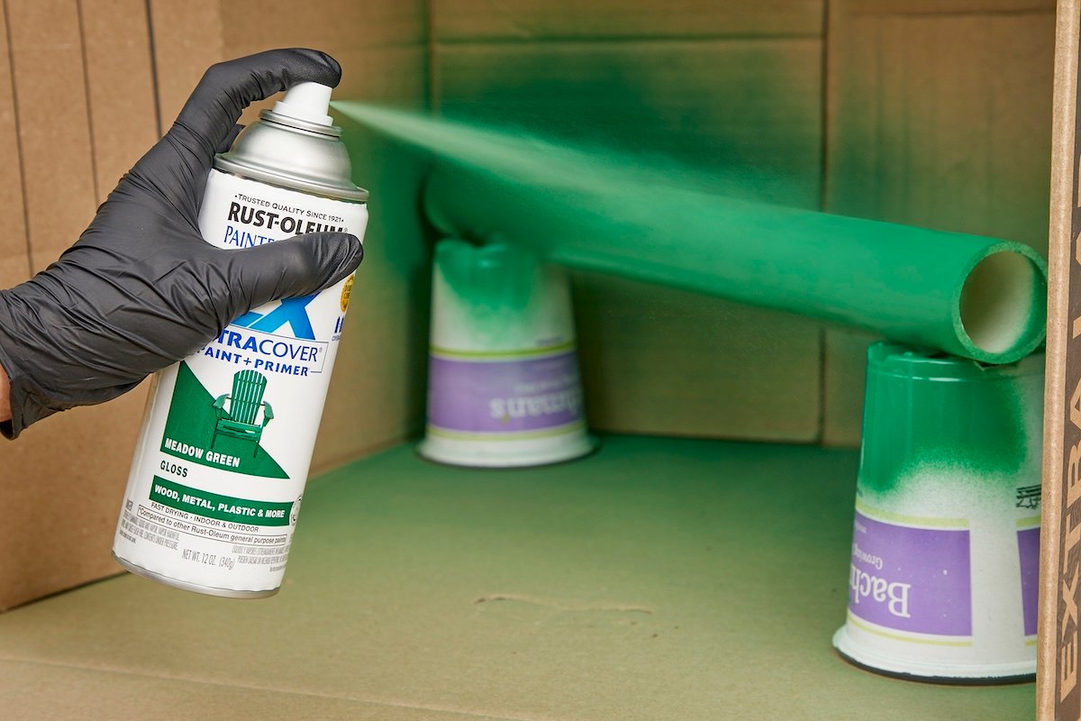 Woman sprays green spray paint on a piece of PVC pipe propped inside a cardboard box.