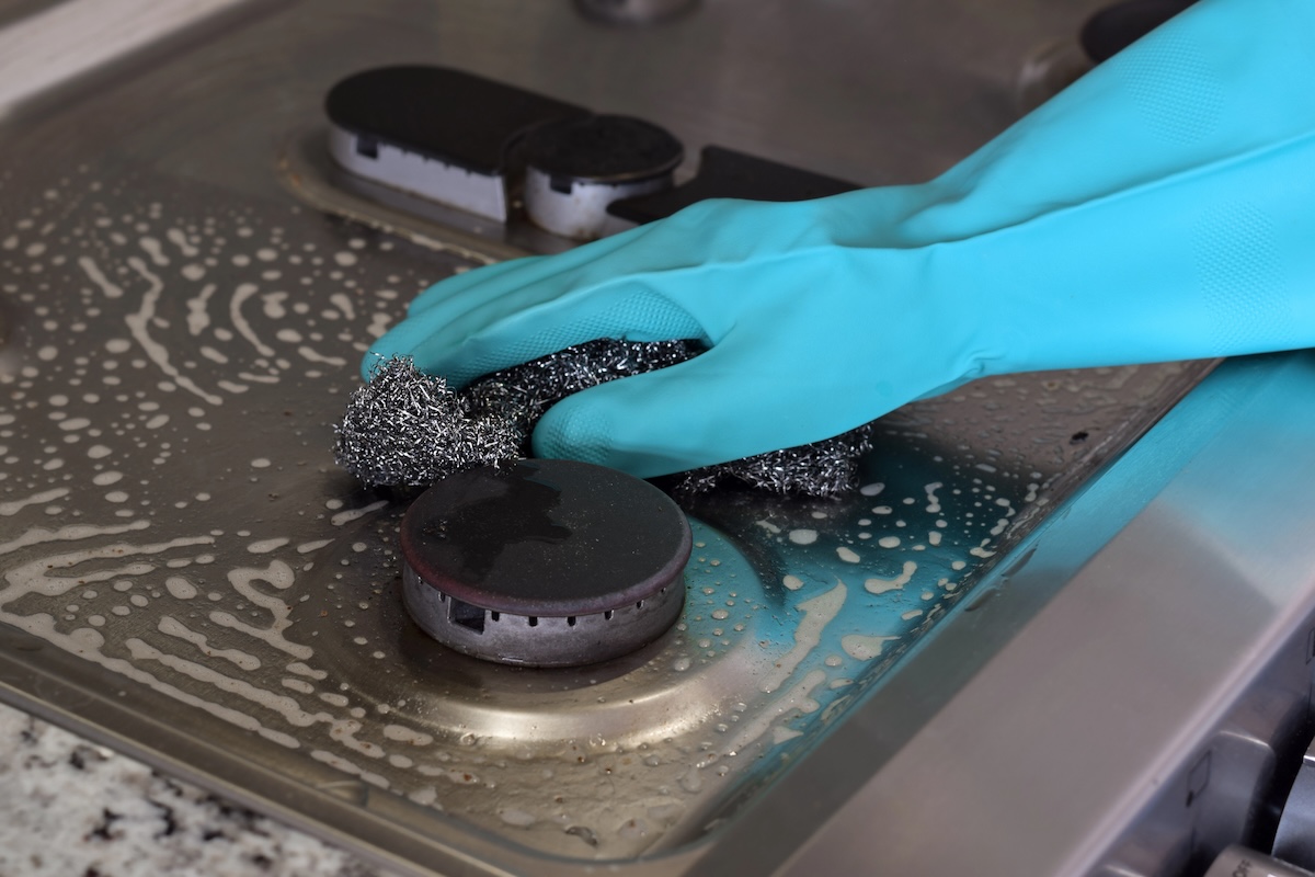 A person wearing a blue rubber glove scrubs a stainless steel stove with steel wool. 
