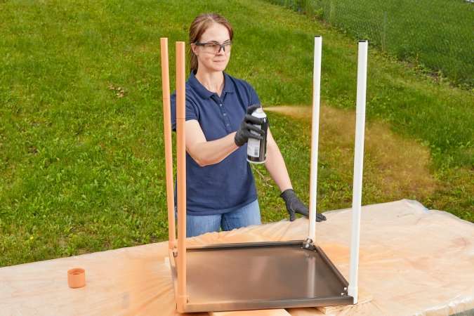 Woman wearing safety glasses spray paints a small metal table outdoors.