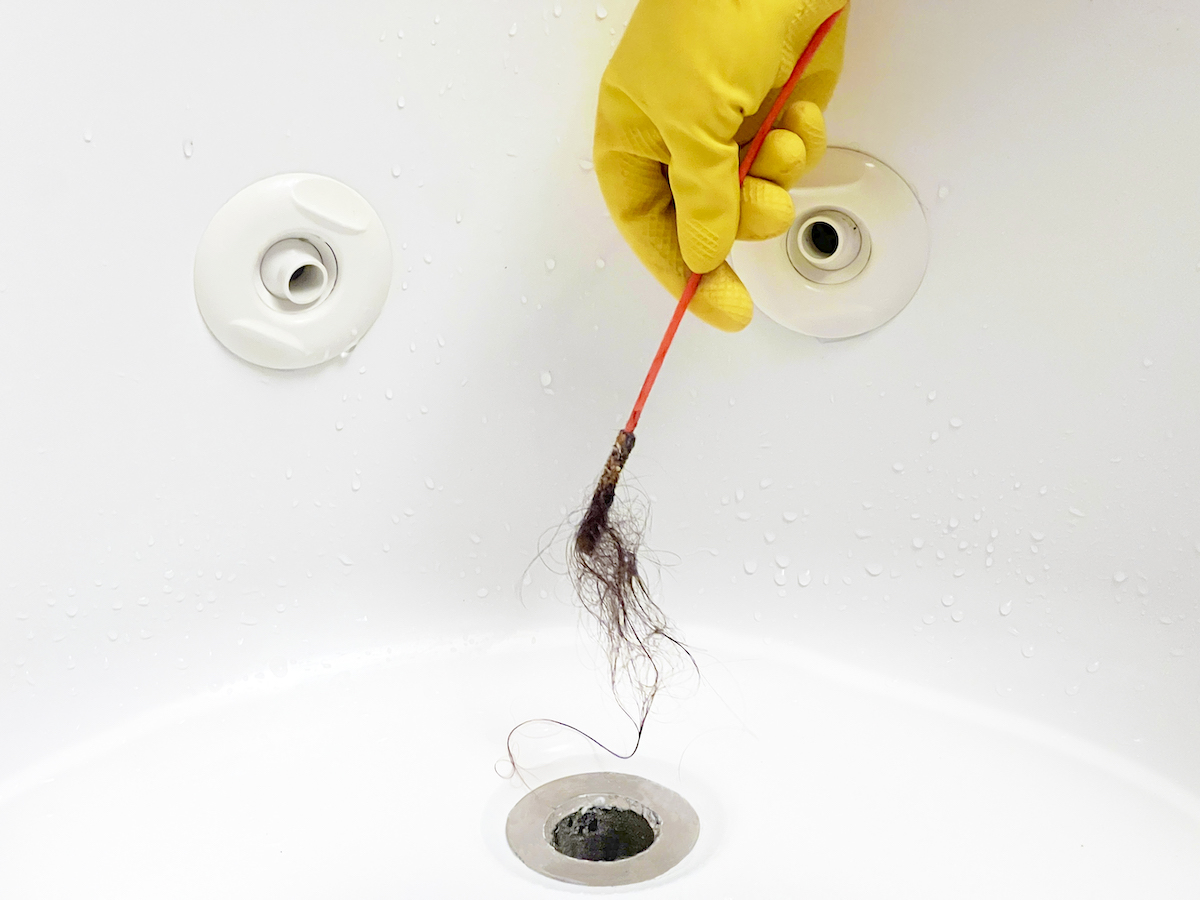 Woman with rubber gloves on uses drain snake to clear tub drain.