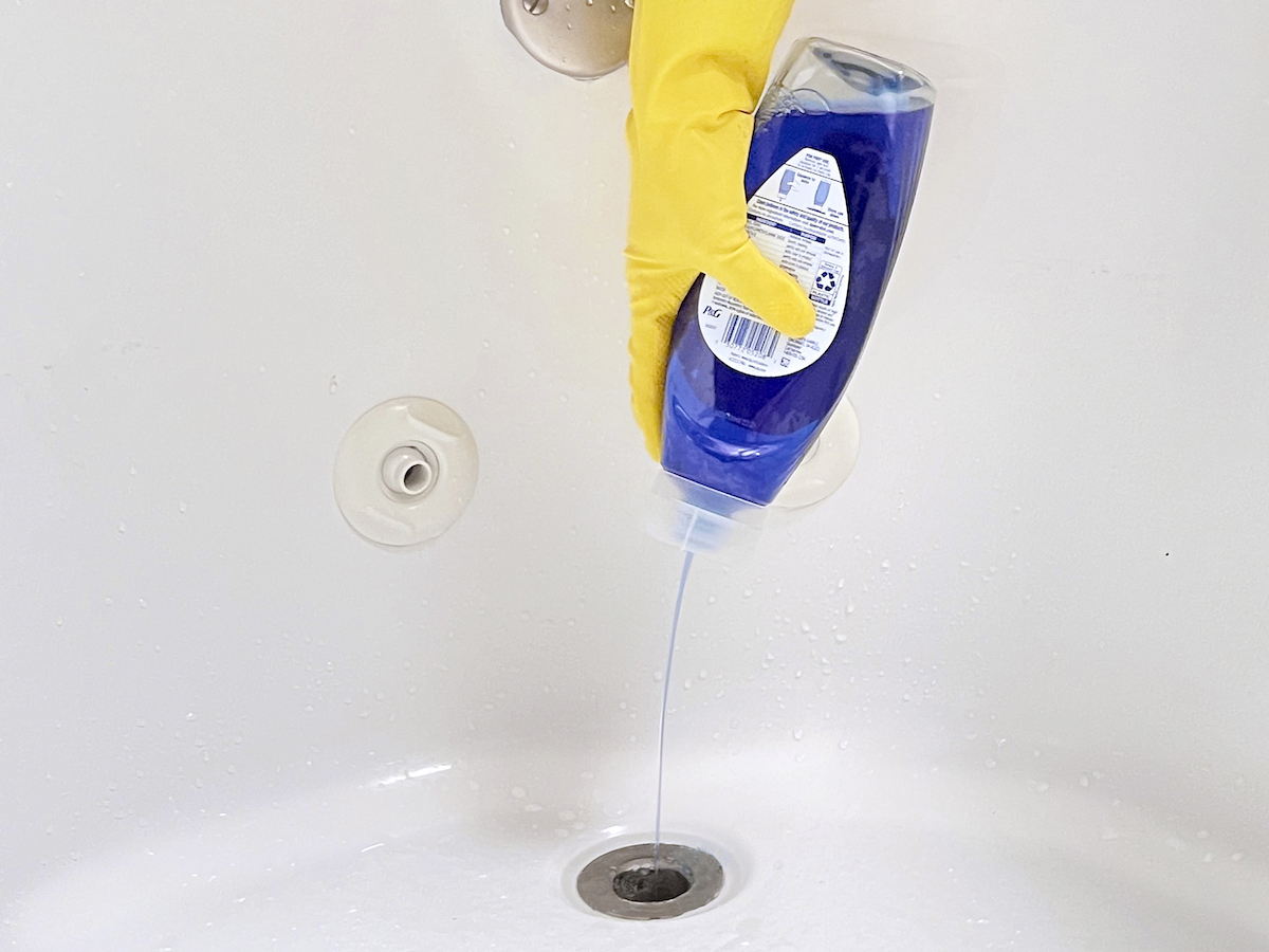 Woman wearing yellow cleaning gloves squirts dish soap into a tub drain.