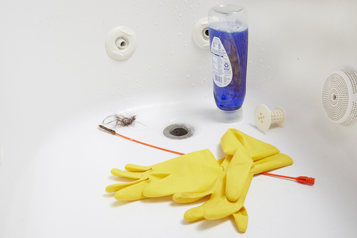 Rubber gloves, drain unclogger, and dish soap inside a white bathtub.