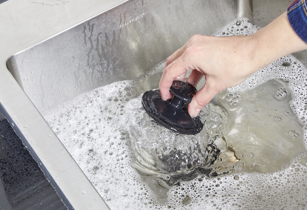 Person pulls sink stopper out of stainless kitchen sink filled with soapy water.