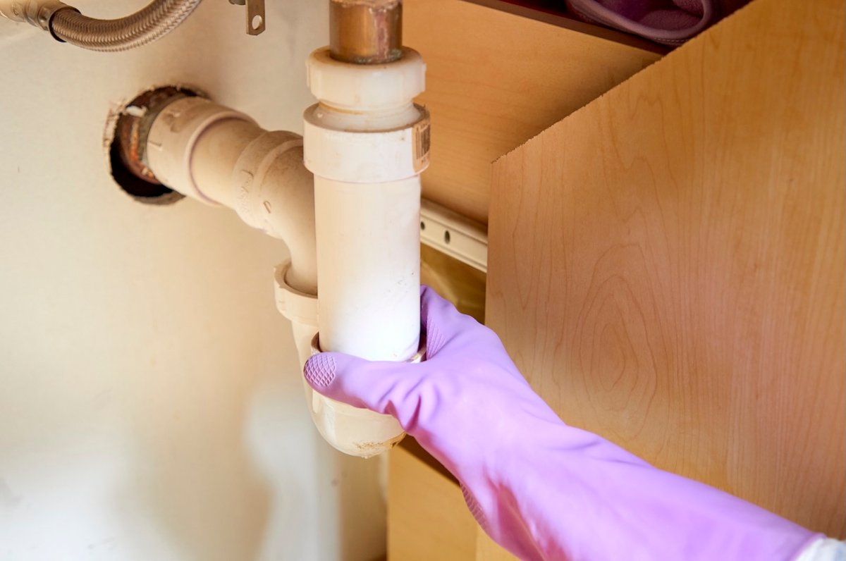 Person wearing purple rubber gloves puts hand on P-trap piping under a sink.