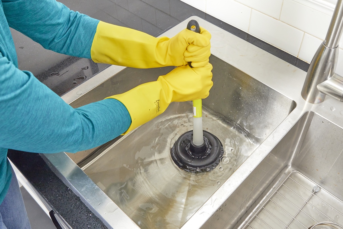 Woman wearing rubber gloves plunges drain on stainless steel kitchen sink.