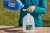 A woman measures blue dish detergent into a measuring spoon before pouring it into a jug of water.