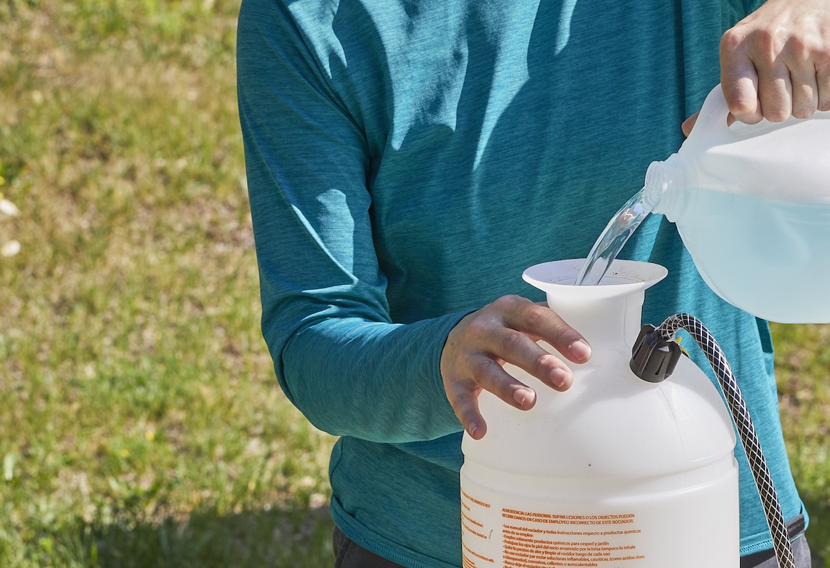 A woman fills a pump sprayer with water from a 1-gallon jug outside.