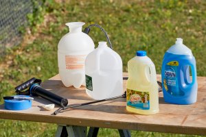 Pump sprayer, cooking oil, dish soap, water jug, and other materials on a table outdoors.
