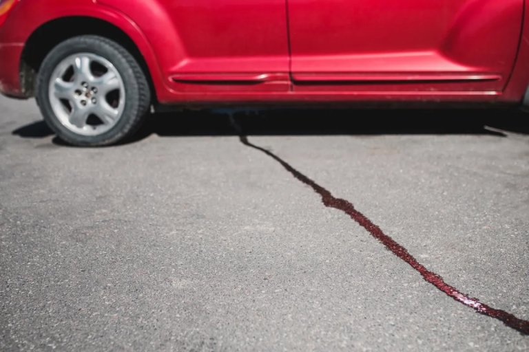 Liquid leaks from under a red car parked on a concrete drive.