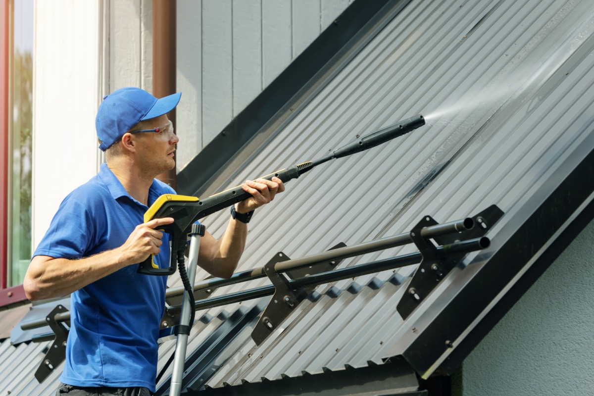 Man wearing a blue shirt and cap pressure washes a roof.