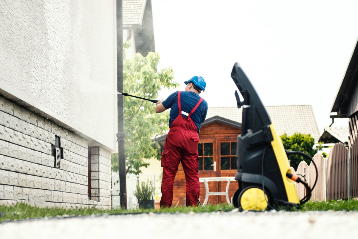 Man wearing red overalls pressure washes an ivory stucco house with the pressure washer in the foreground.