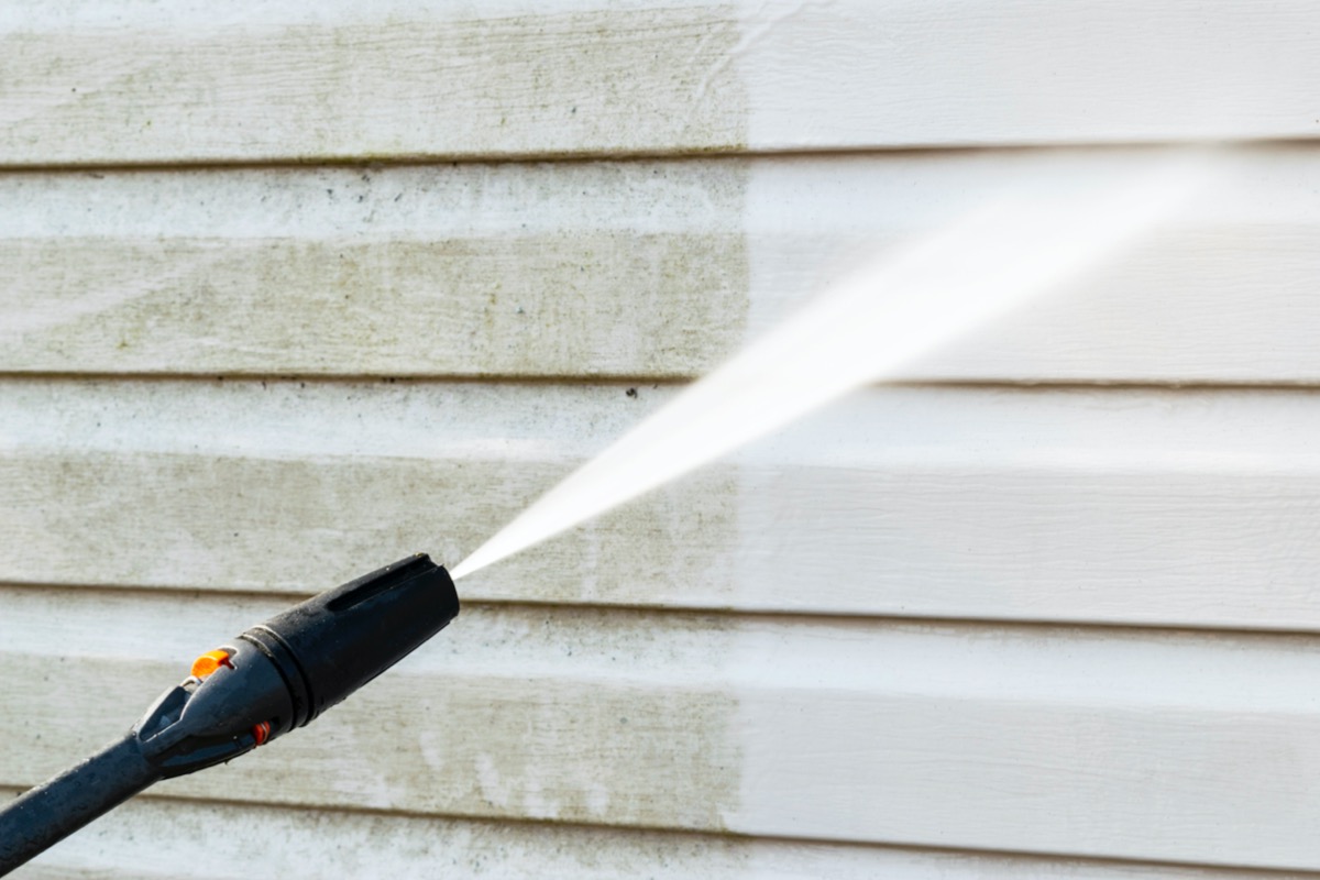 A close up of a pressure washer nozzle cleaning grey siding on a house.