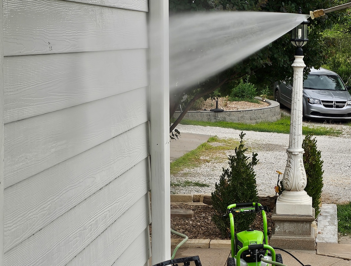 Pressure washer nozzle sprays water at grey house siding, the pressure washer in the background.