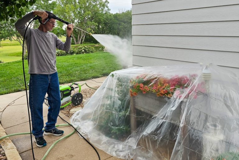 Man pressure washes siding of house, plants underneath covered with plastic.