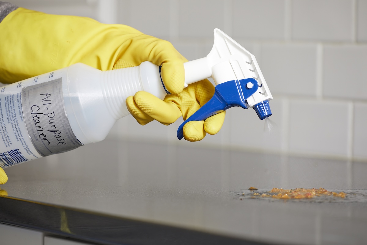 Person wearing yellow rubber gloves points a spray bottle labeled 'all-purpose cleaner' at a mess on the countertop.