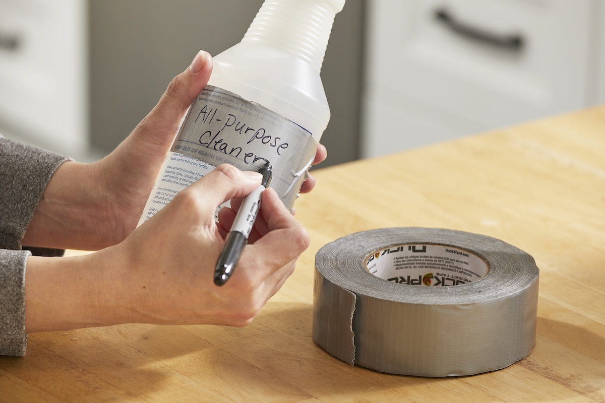 Woman labels a spray bottle "all-purpose cleaner" written in marker on duck tape.