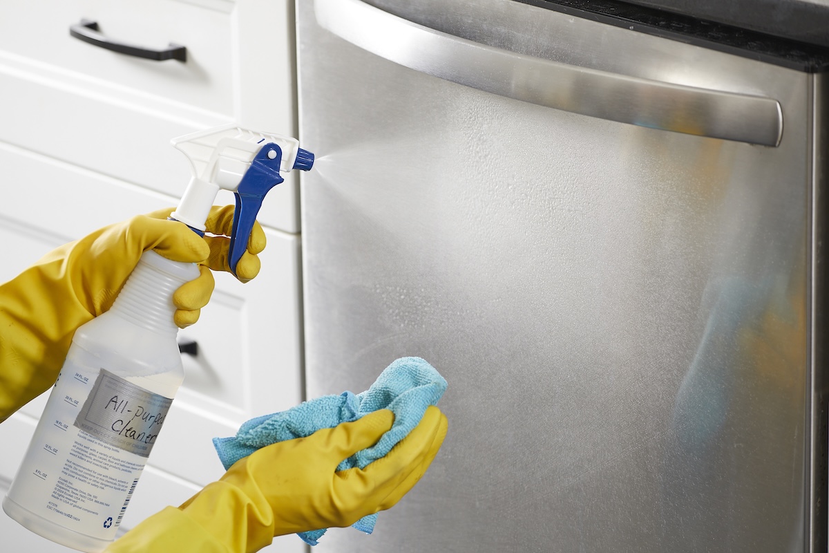 Person wearing yellow rubber gloves sprays a dishwasher with homemade cleaner, holding microfiber cloth in right hand.