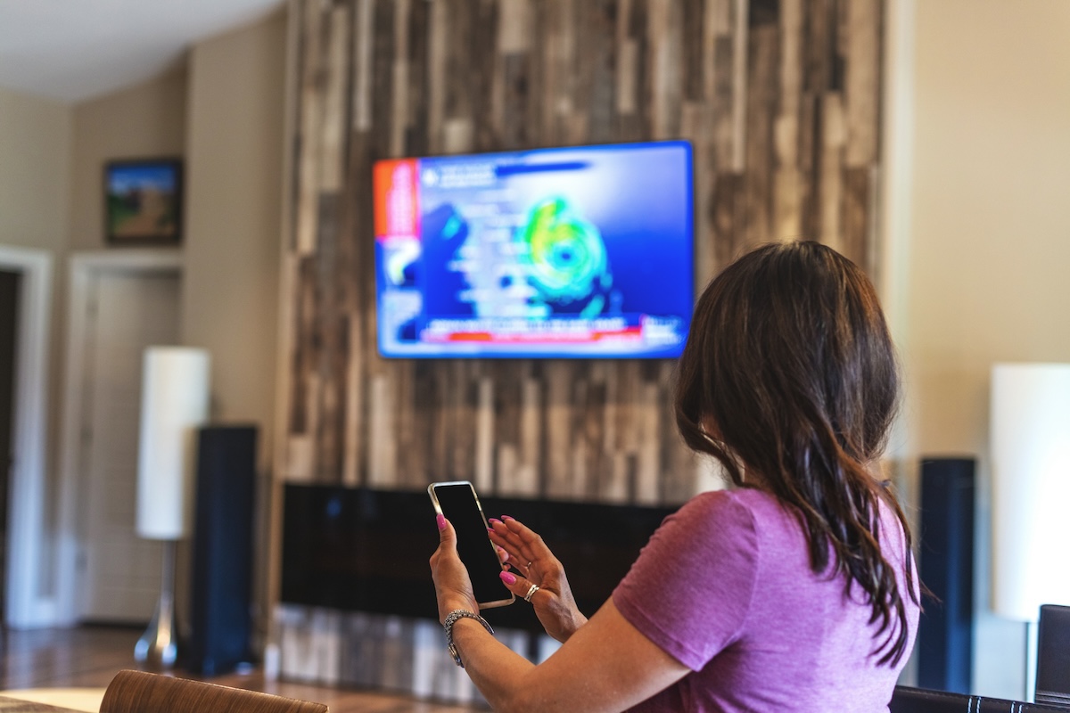 A woman watches a hurricane forecast on the television and reaches for her cell phone.