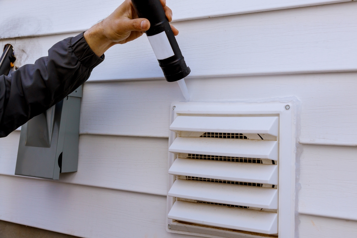 A person’s arm is seen using caulk to seal the exterior vent to a home.