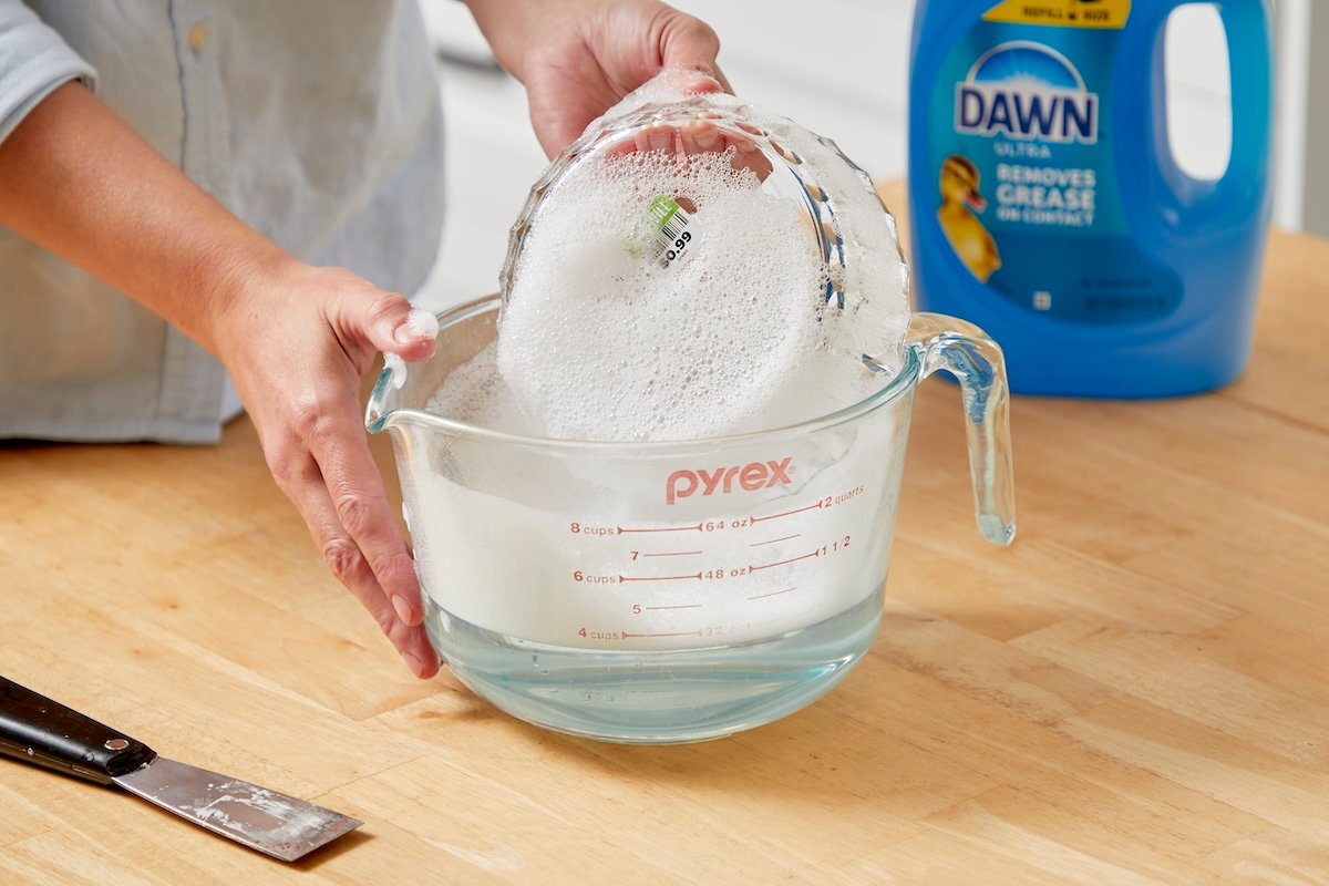 Woman washes a glass dish with a sticker in a large measuring cup with soapy dish water.