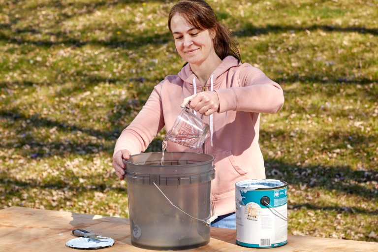 Woman pours a cup of water into a large bucket, a can of white paint next to the bucket.