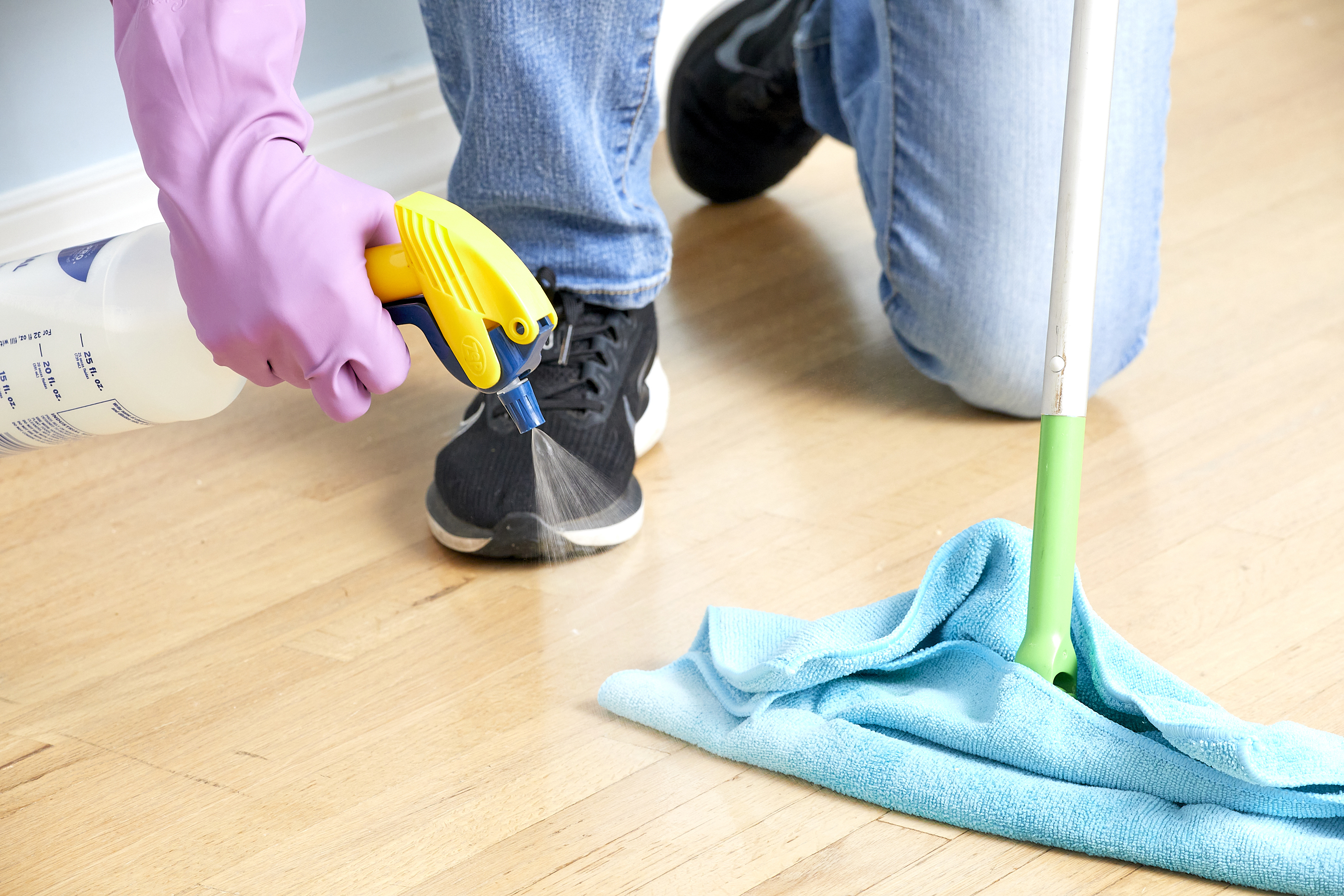 Woman sprays cleaner onto wood floor, a microfiber mop nearby for mopping.