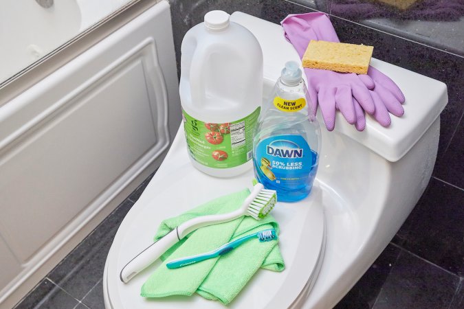 Materials needed to clean a toilet tank sitting on top of the toilet, including vinegar and dish soap.