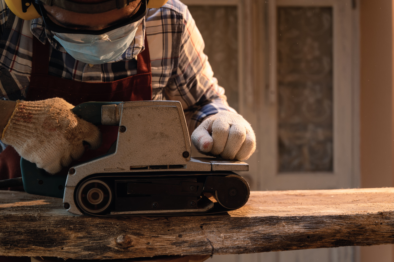 Carpenter use a hand-held electric sanding machine belt sander to level surface wood in carpentry workshop.