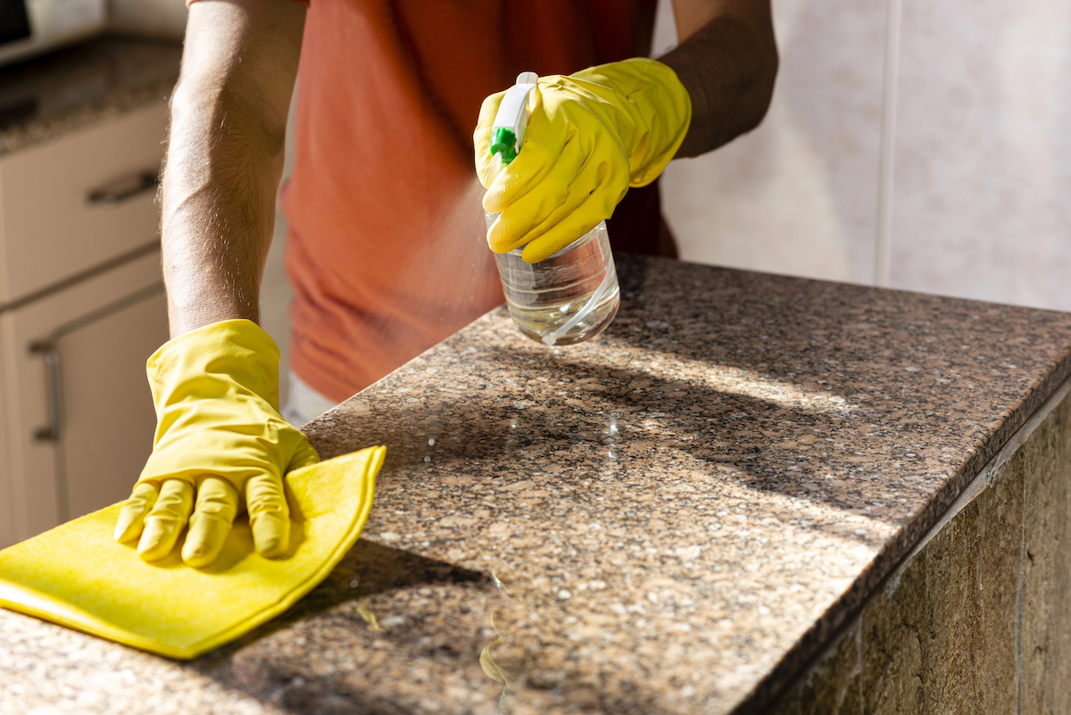 Woman wearing an apron and rubber gloves cleaning granite countertops with a spray bottle and yellow cloth.