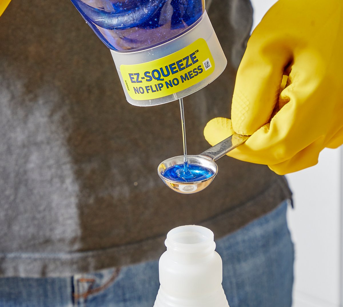 Woman wearing yellow rubber gloves pours dish soap into a measuring spoon above a spray bottle.