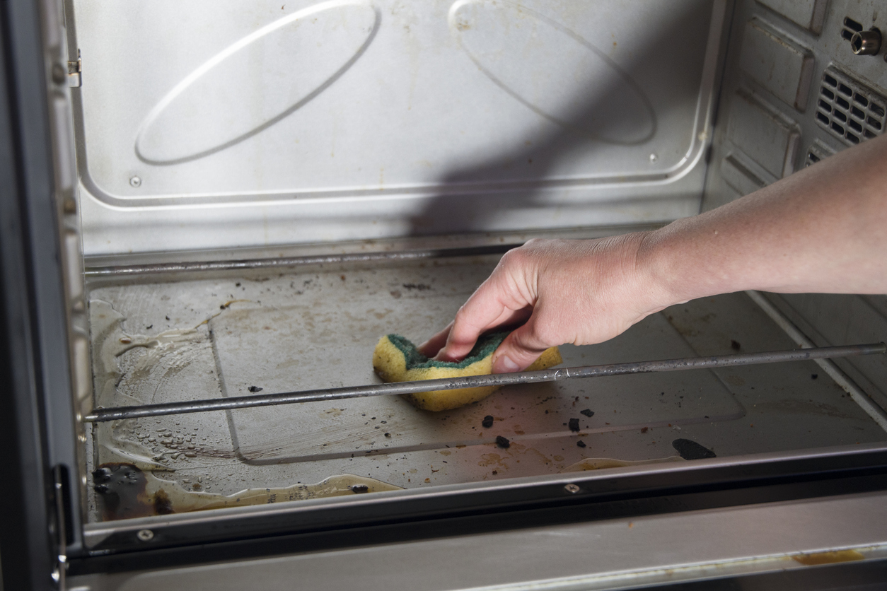 Woman's hand uses yellow and green sponge to clean oven grime before turning on self-cleaning function.